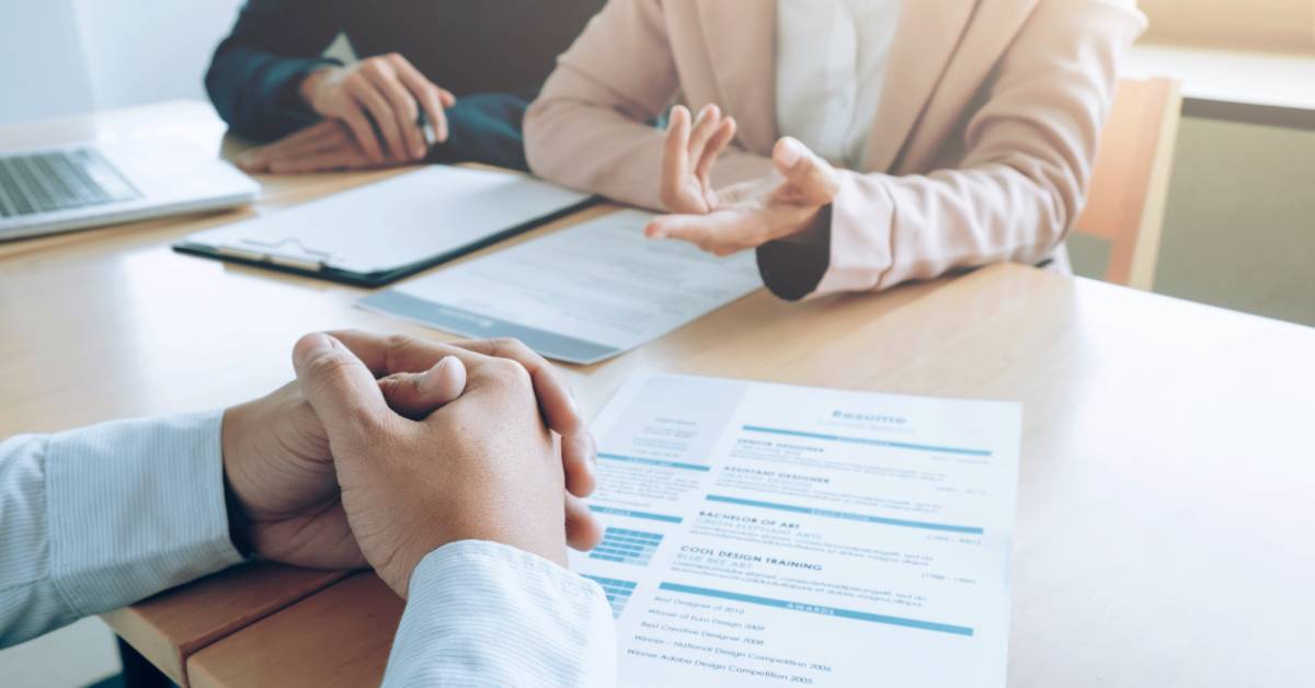 The torso and hands of two people interviewing a person for a job. The interviewee's resume is on the table in front of them.