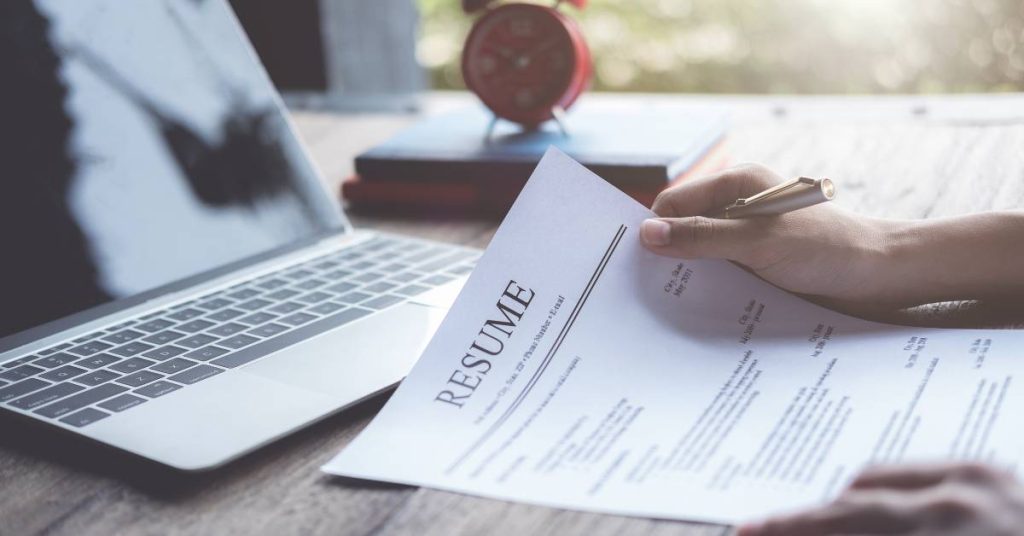 A person holds a pen and a white sheet of paper with "RESUME" printed at the top at a desk in front of a laptop.