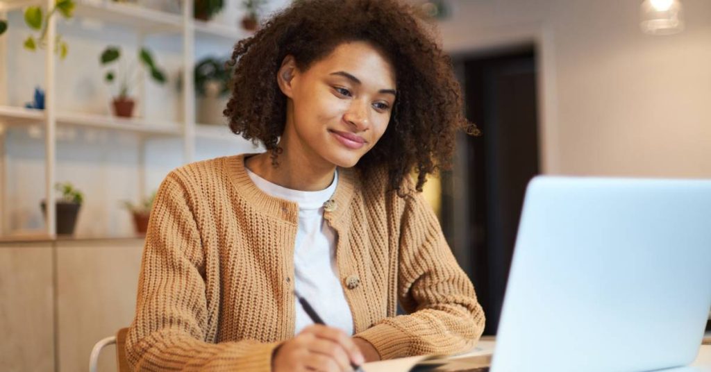 A young, confident woman in a brown sweater takes notes on a notepad at a desk in front of a laptop computer.