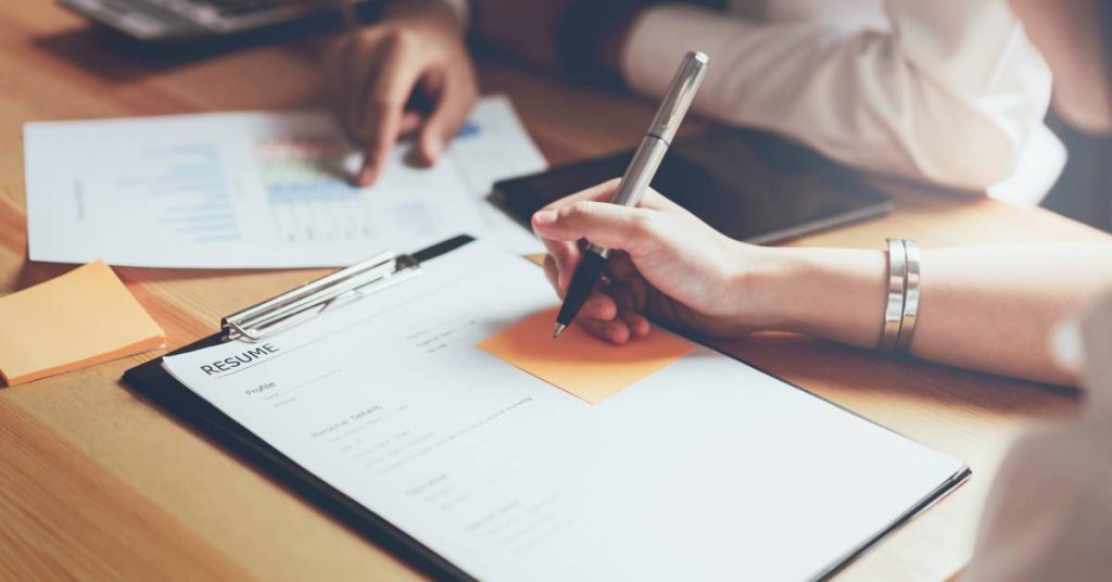 Two pairs of hands, one writing with pen on a sticky note on a clipboard while another points to statistics on another page.