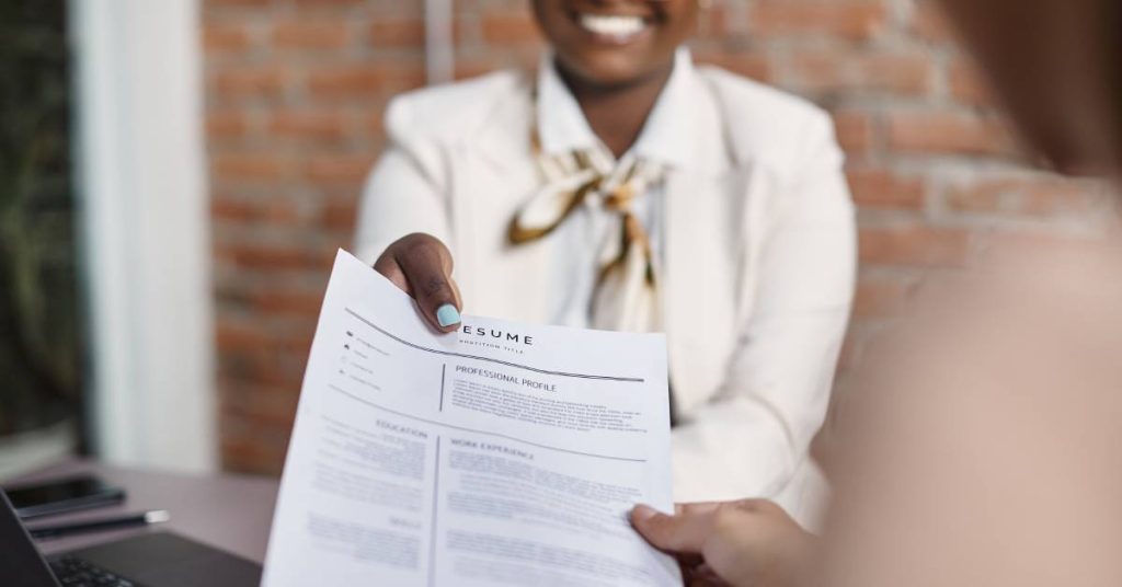 A point-of-view of an interviewer being handed a professional resume from a young woman in business attire.